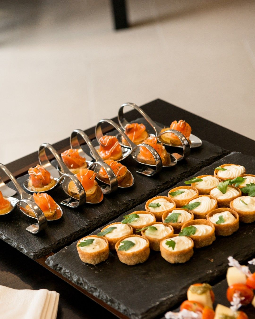 Cured salmon canapés on spoons, beside cream-filled tartlets on slate.