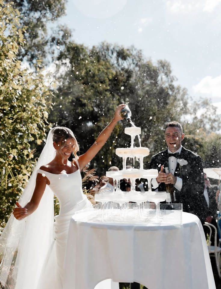 A bride pouring champagne over a tower of coupes in the garden while the groom laughs.