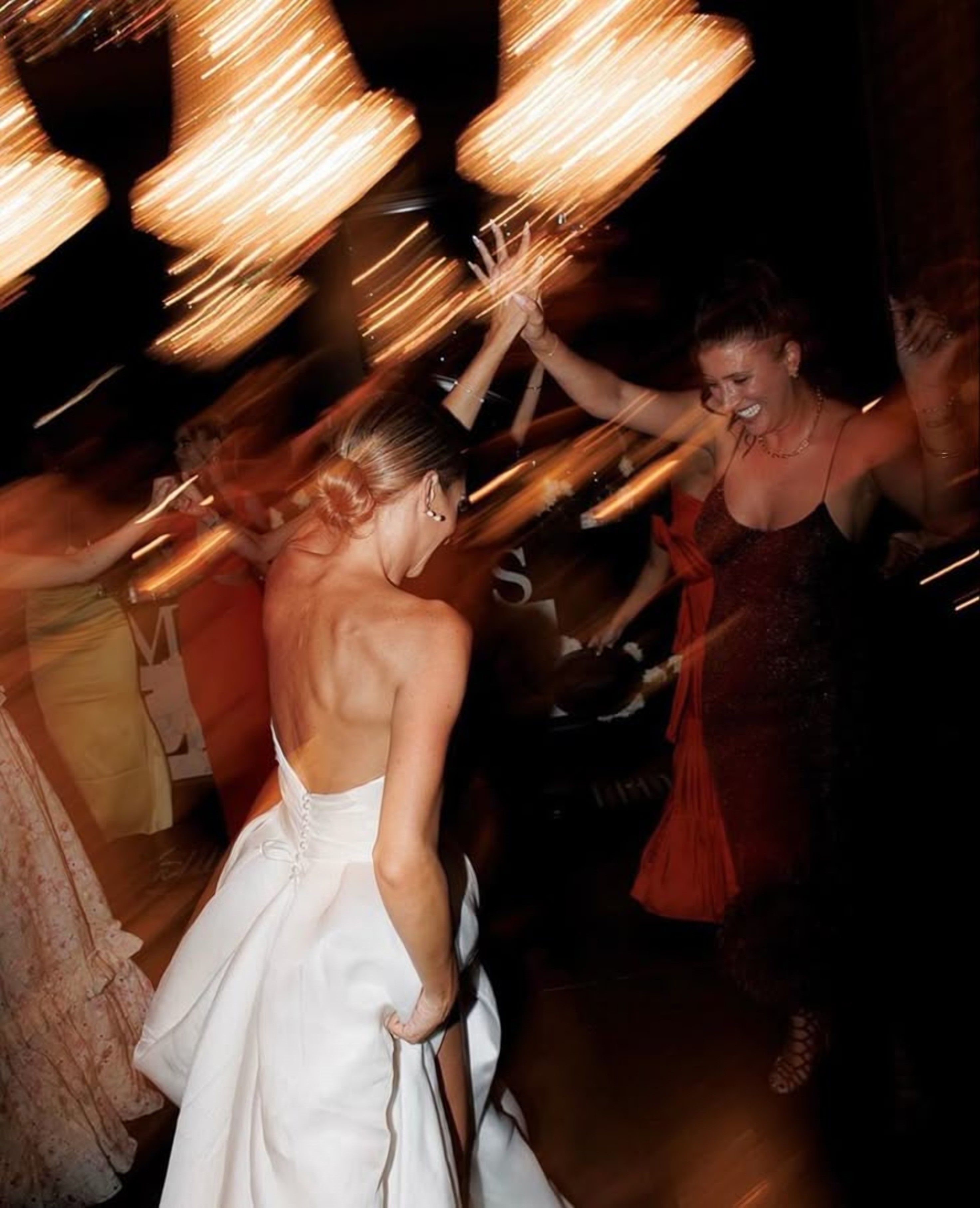 A bride dancing with friends under chandeliers, the light blurred with movement.