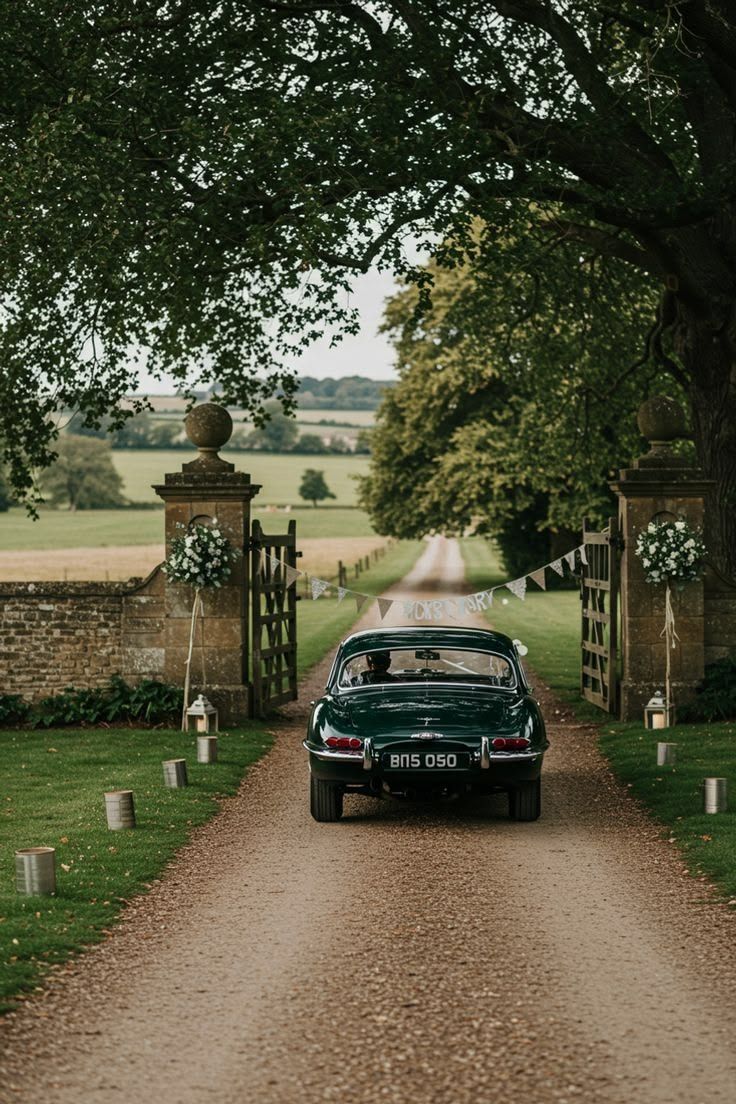 A green vintage car driving through stone gate piers dressed with white flowers, onto a tree-lined drive across open fields.