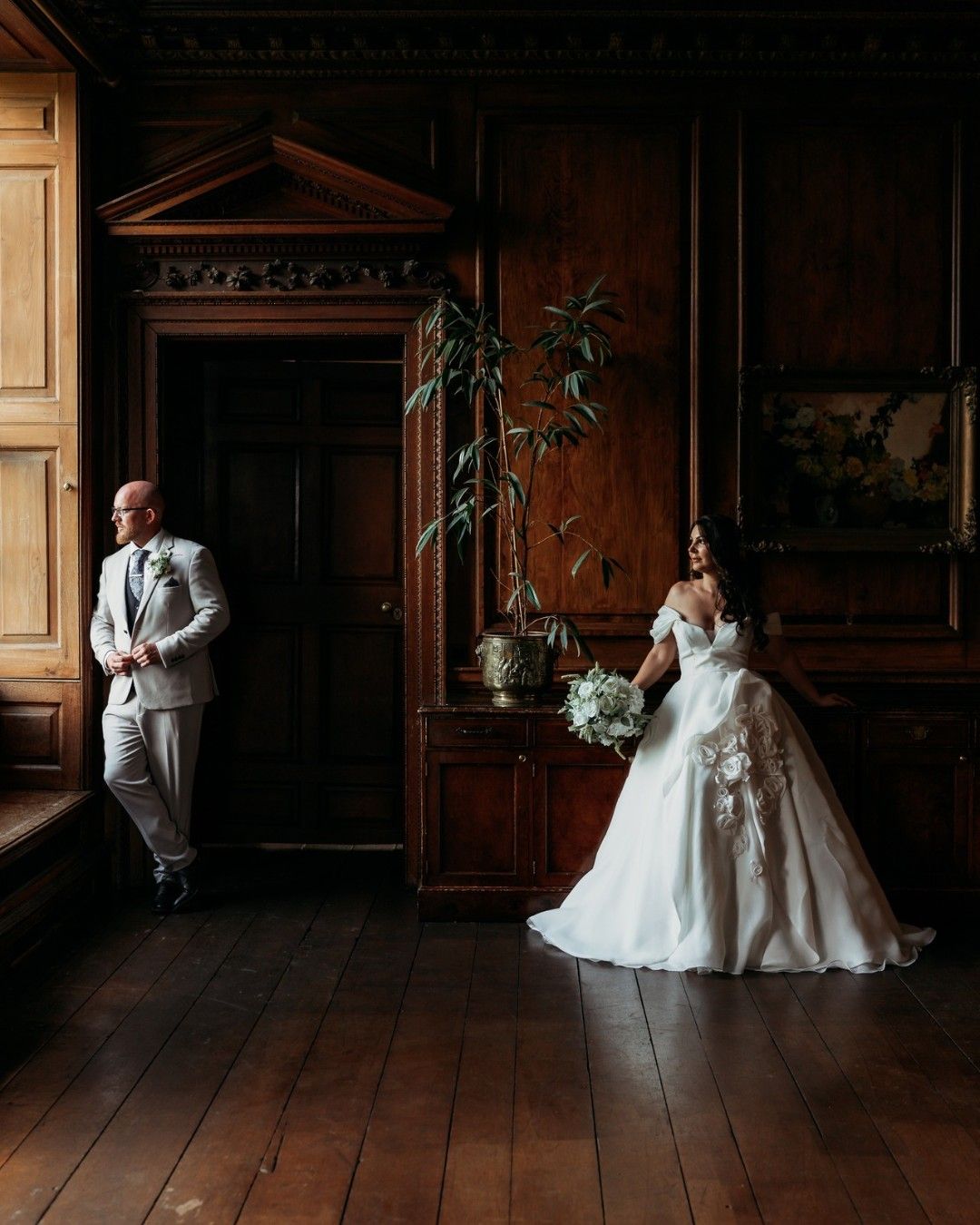 A couple in a wood-panelled room, light falling through a tall window.