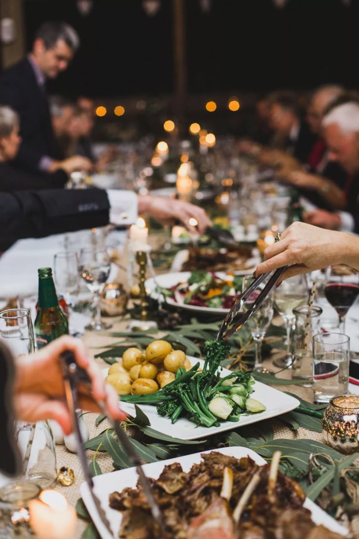 Guests passing sharing platters of potatoes, greens and lamb along a long candlelit table.