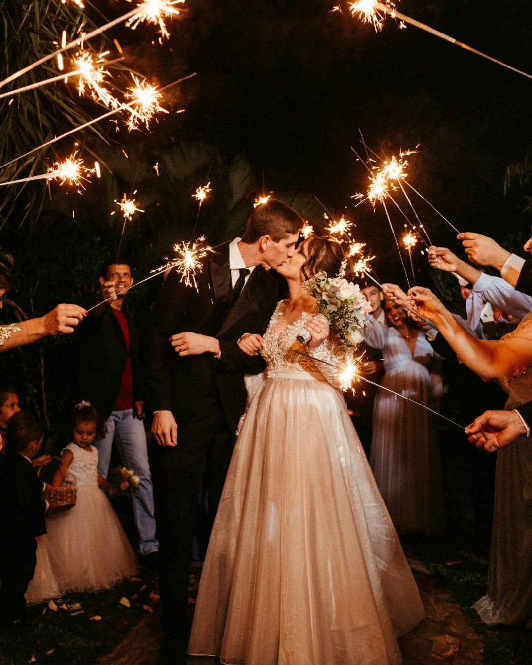 A couple kissing in a tunnel of sparklers held by their guests at night.